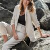 A smiling blonde woman in a cream blazer sits gracefully on large grey rocks outdoors in daylight.