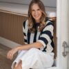 A smiling blonde woman wearing a striped sweater sits comfortably in a bright, modern, coastal-inspired room.