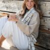 A smiling blonde woman wearing a cozy beige sweater sits relaxed against a weathered wooden beach wall.
