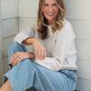 A smiling blonde woman sits against a bright wooden wall, wearing casual jeans and a cream sweater.