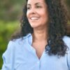 A smiling woman with long curly hair wears a blue striped shirt in a natural outdoor setting.