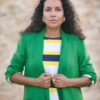 A woman with curly hair wearing a bright green blazer and striped top poses against a stone wall.