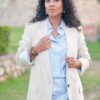 A woman with curly hair wears a light blazer and striped shirt in a soft garden setting.