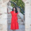 A woman with curly dark hair wearing a bright red dress stands inside a stone archway outdoors.