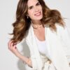 rachael-warner-1 A smiling woman with wavy brunette hair wears a white blazer against a clean bright studio background.