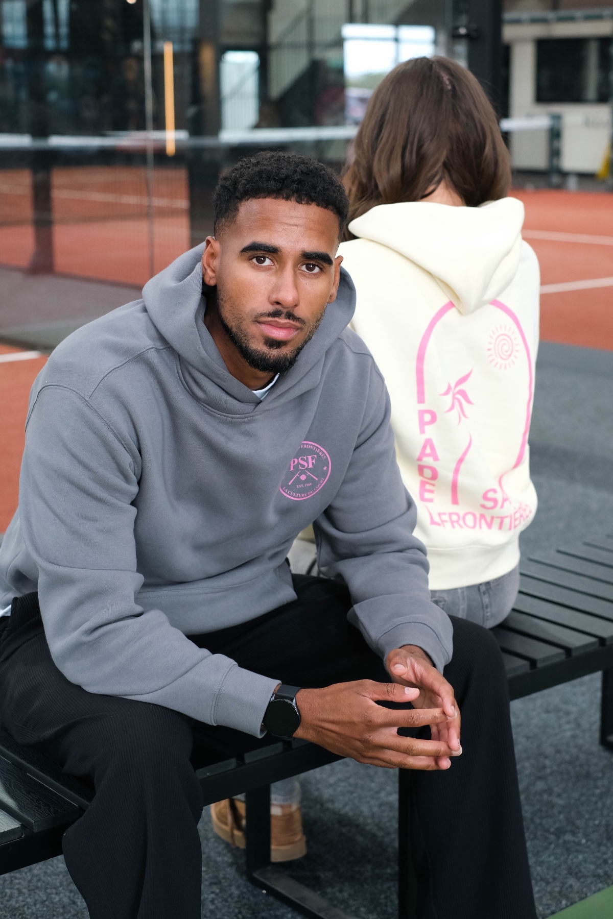 A man in a grey hoodie sits on a bench with a partner at a padel court.