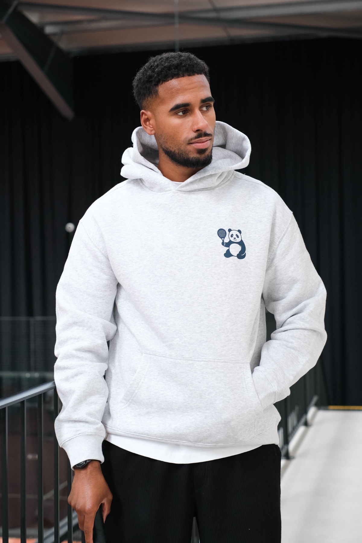 A handsome man wearing a grey panda-logo hoodie looks away while standing in a bright indoor studio.