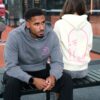 A man in a grey hoodie sits on a bench beside a woman wearing a cream sweatshirt.