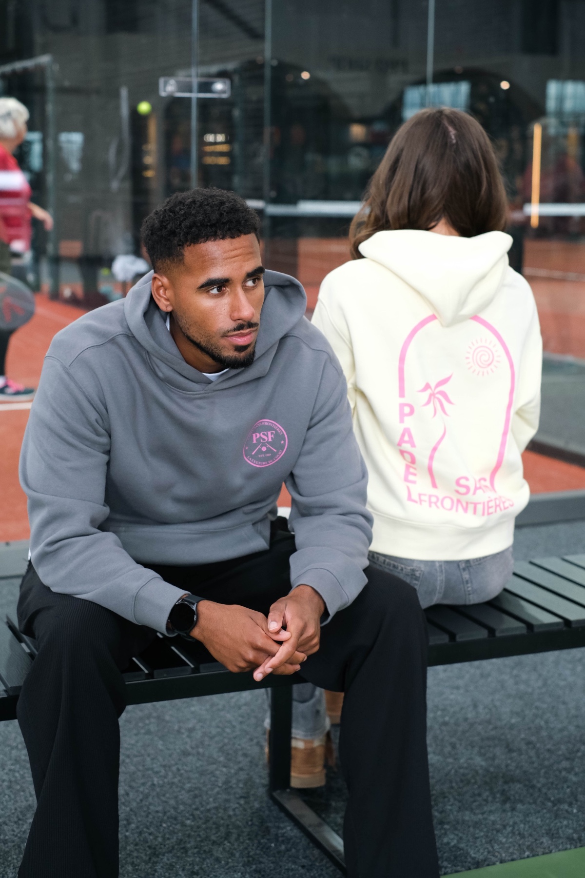 A man in a grey hoodie sits on a bench beside a woman wearing a cream sweatshirt.