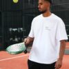 A man in a white t-shirt holds a padel racket on an indoor court during a game.