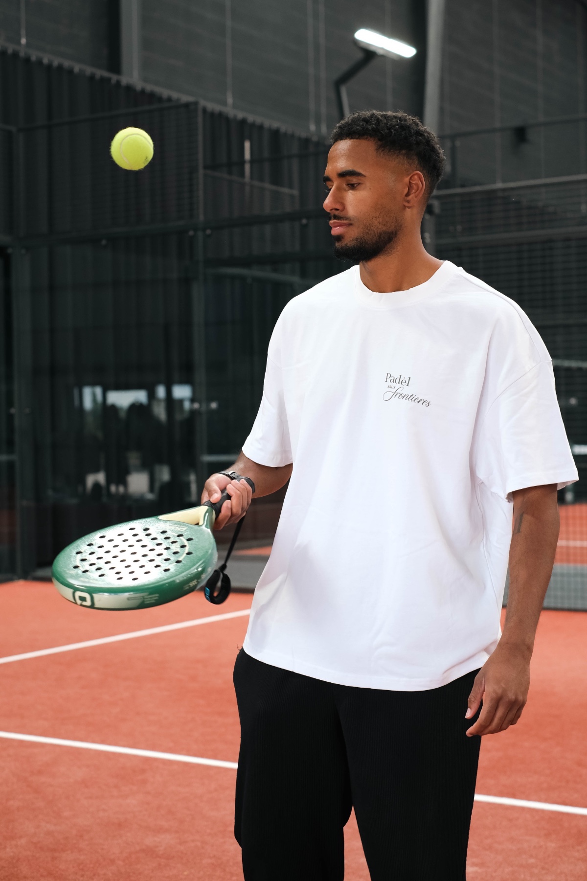 A man in a white t-shirt holds a padel racket on an indoor court during a game.