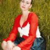 A woman in a red and white blouse sits in a field of tall green grass outdoors.