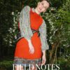 A stylish model wears layered autumn-inspired clothing while posing outdoors in front of white flowering bushes.