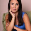 A warm, natural-looking brunette woman in a blue top sitting in a chair against a pink backdrop.