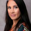 A woman with long dark hair and subtle grey roots poses for a clean professional studio headshot.