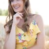 A smiling woman in a vibrant floral yellow dress stands in a sunny, soft-focus outdoor golden field.