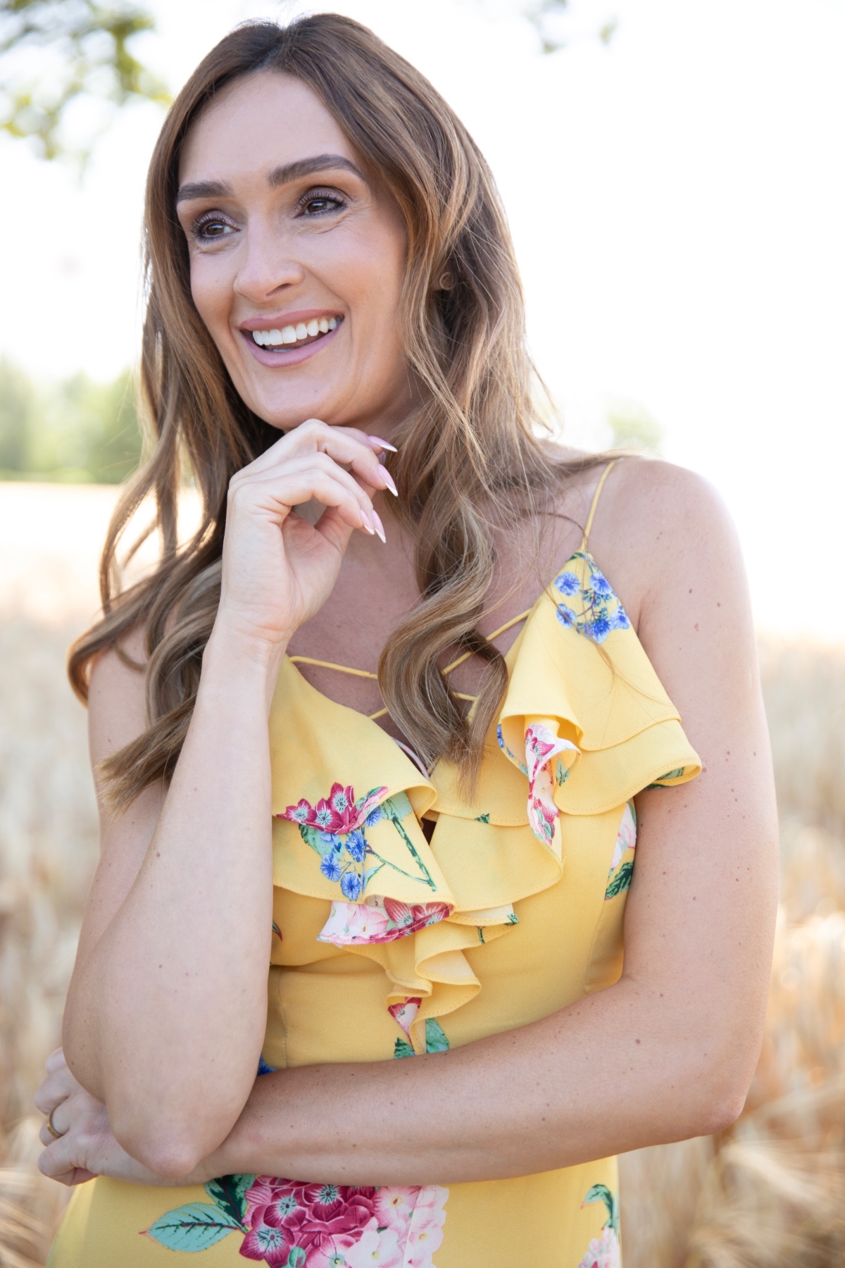 A smiling woman in a vibrant floral yellow dress stands in a sunny, soft-focus outdoor golden field.