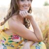 A smiling woman in a vibrant floral summer dress poses naturally in a sunny outdoor wheat field.