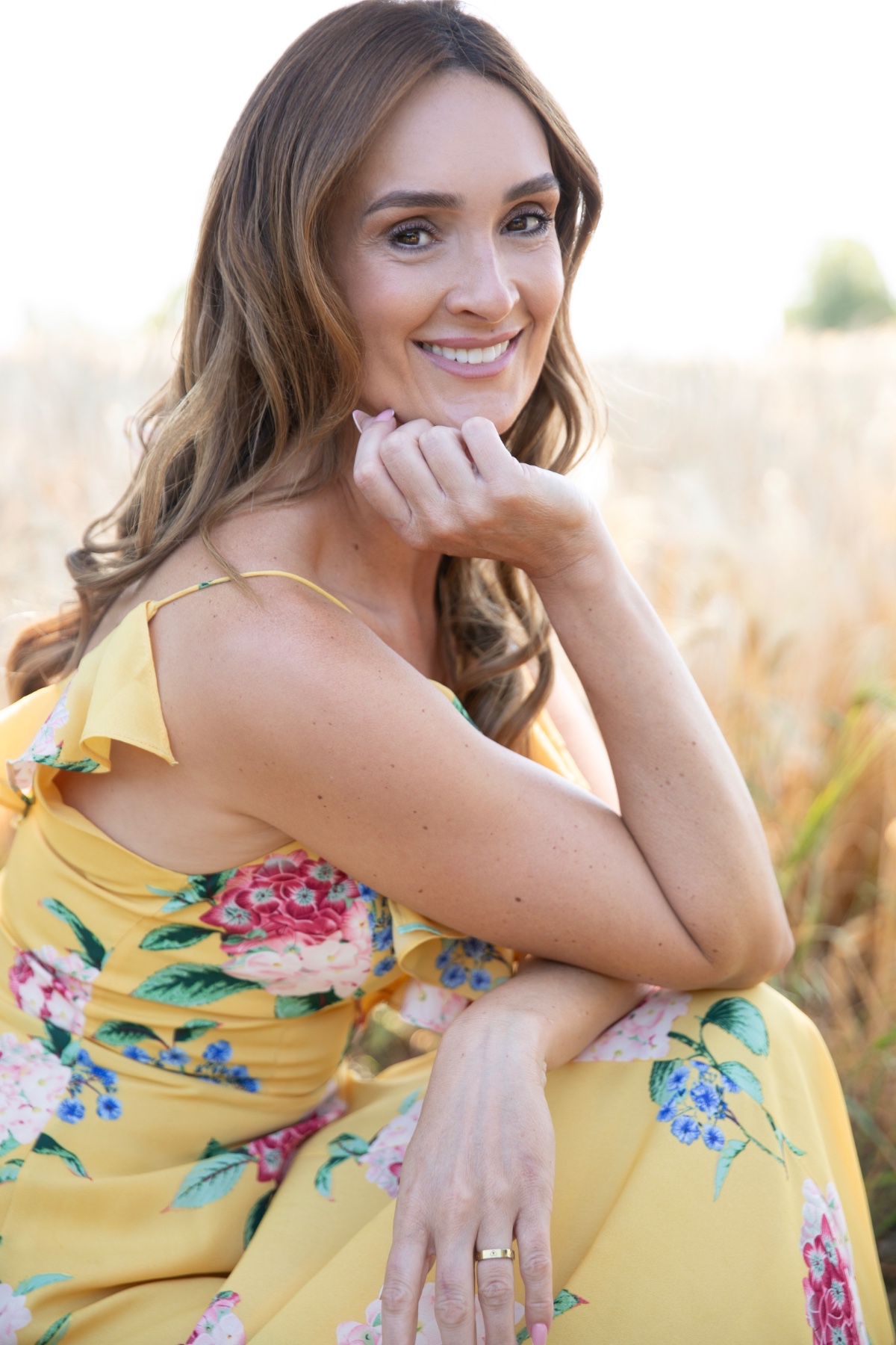 A smiling woman in a vibrant floral summer dress poses naturally in a sunny outdoor wheat field.