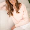 A smiling woman with wavy brunette hair sits on a white couch wearing a soft pink sweater.
