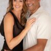 A smiling couple standing close together, dressed in smart-casual attire against a soft neutral textured background.