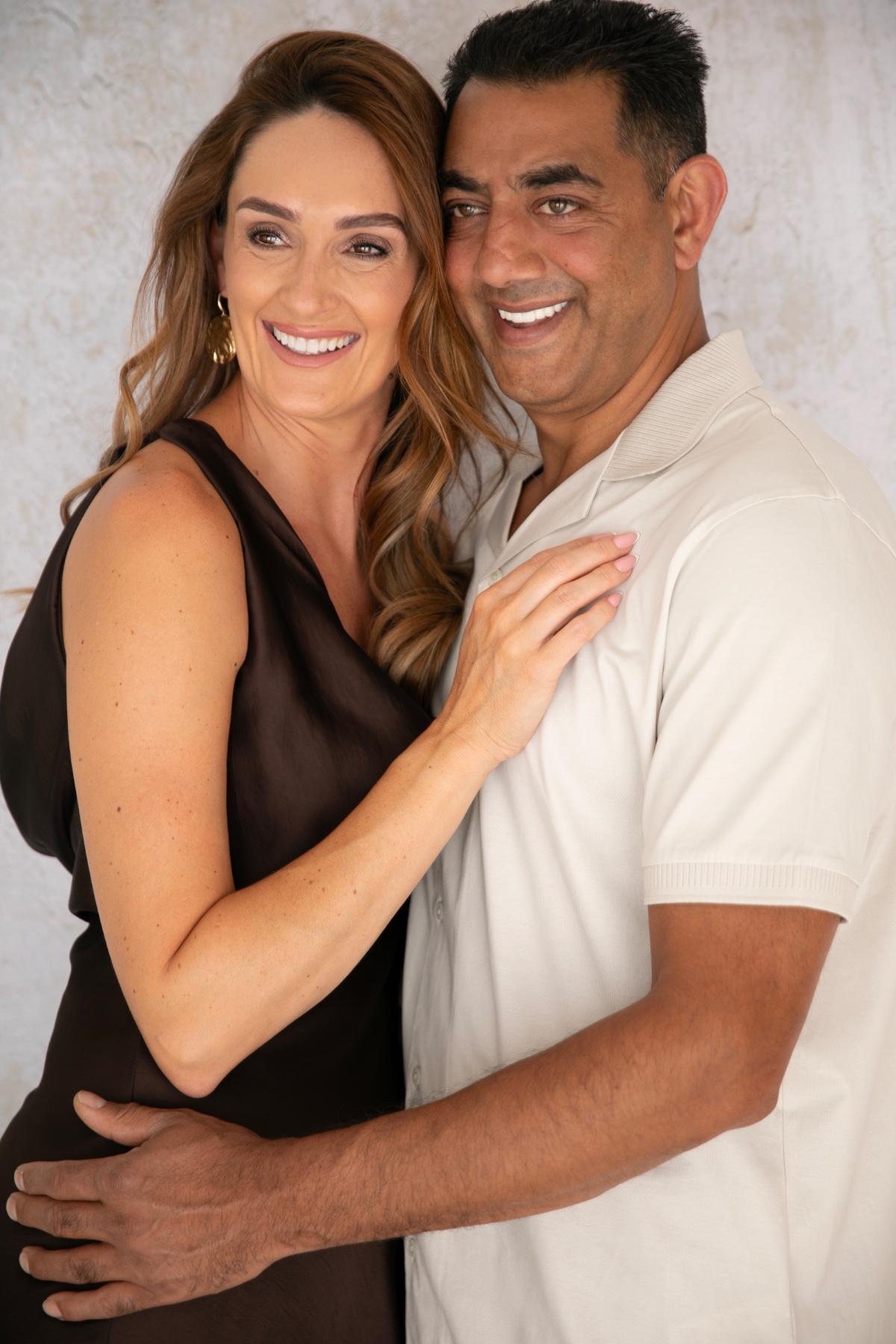 A smiling couple standing close together, dressed in smart-casual attire against a soft neutral textured background.