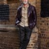 peter-williams-13 A silver-haired mature man in a leather jacket leans against a textured brick wall outdoors.