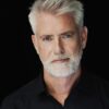peter-williams-7 A striking portrait of a middle-aged man with silver hair and a beard against a black background.