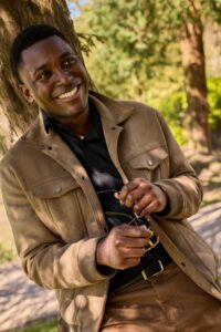 A smiling man holding sunglasses leans against a tree in a sunlit, natural outdoor park setting.