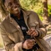 deondre-cumberbatch-11 A smiling man holding sunglasses leans against a tree in a sunlit, natural outdoor park setting.