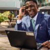 deondre-cumberbatch-14 A smiling professional man in a suit works on his laptop at an outdoor plaza cafe table.