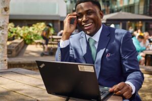 A smiling professional man in a suit works on his laptop at an outdoor plaza cafe table.