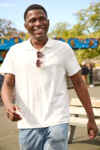 A smiling man in a white polo shirt walks through a sunny, outdoor amusement park setting.