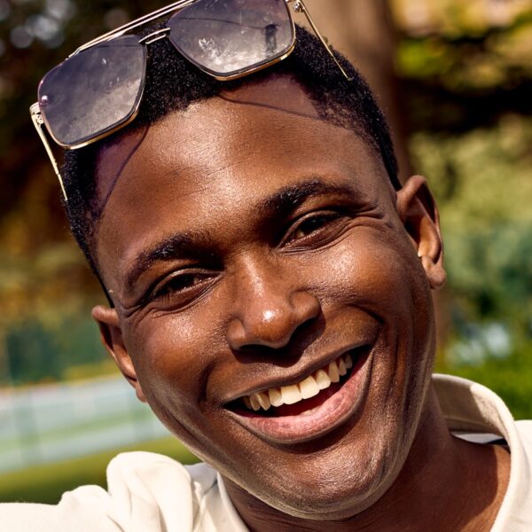 deondre-cumberbatch-1 A smiling man wearing a white polo shirt and sunglasses sits outdoors in the bright summer sunshine.