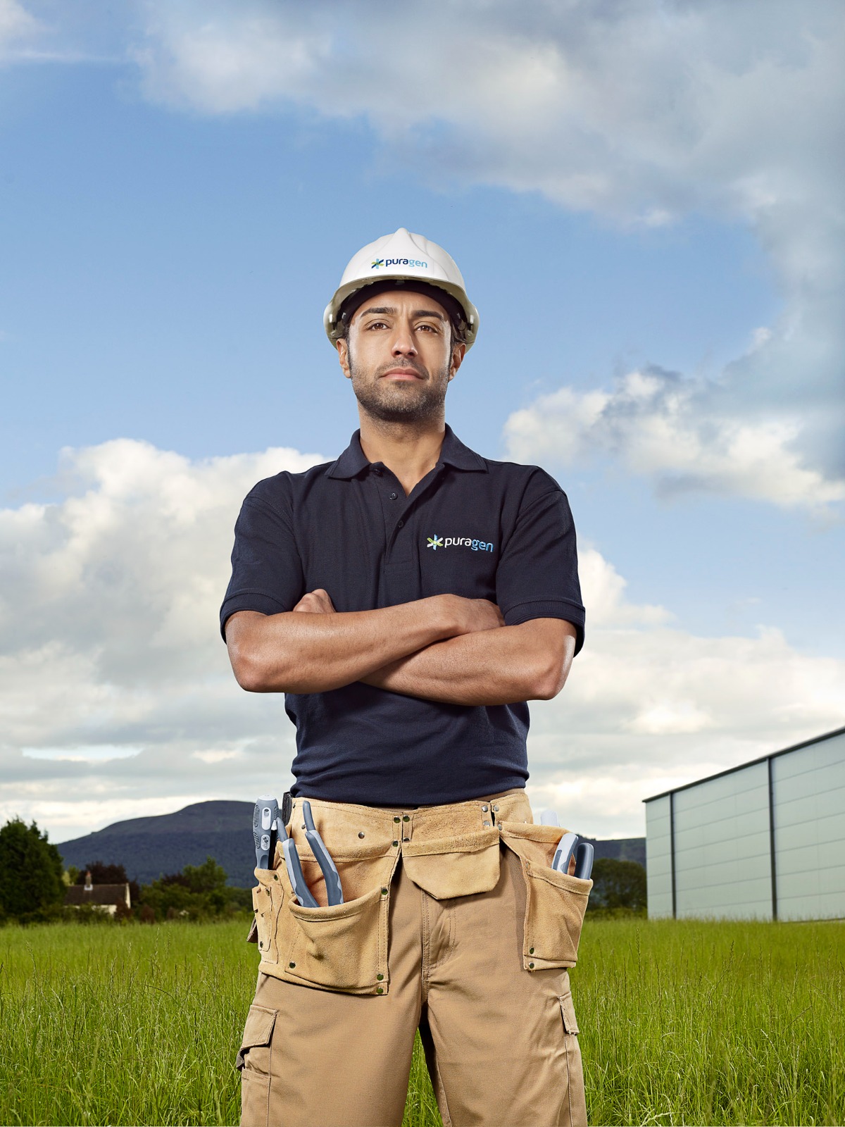 A male worker wearing a branded hard hat and tool belt stands confidently in a grassy field.