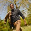 deondre-cumberbatch-12 A smiling man walks through a sunny park holding a jacket over his shoulder, radiating warmth and energy.
