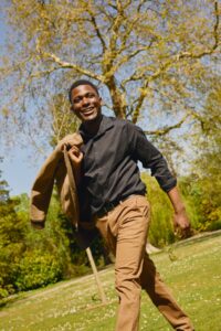A smiling man walks through a sunny park holding a jacket over his shoulder, radiating warmth and energy.