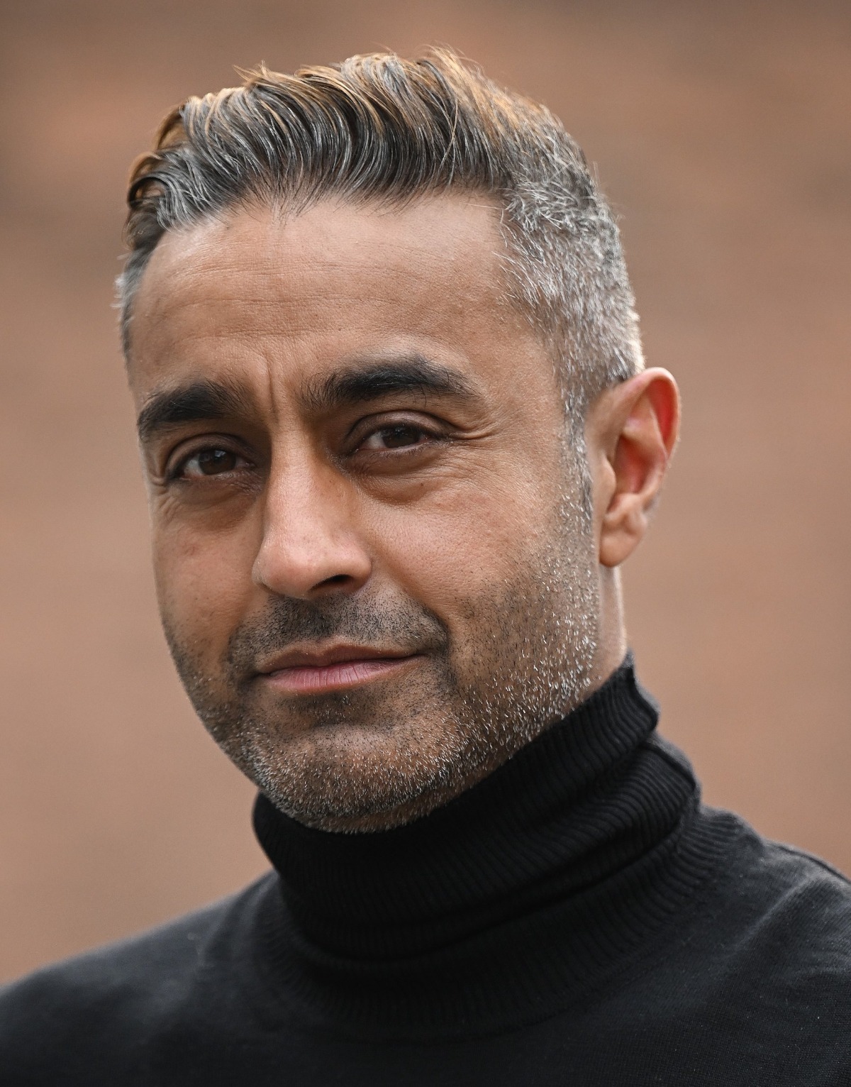 A portrait of a middle-aged man with salt and pepper hair, wearing a black turtleneck jumper outdoors.