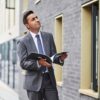 mohan-randhawa-7 A professional man in a grey suit holding a brochure stands outdoors near a modern brick building.