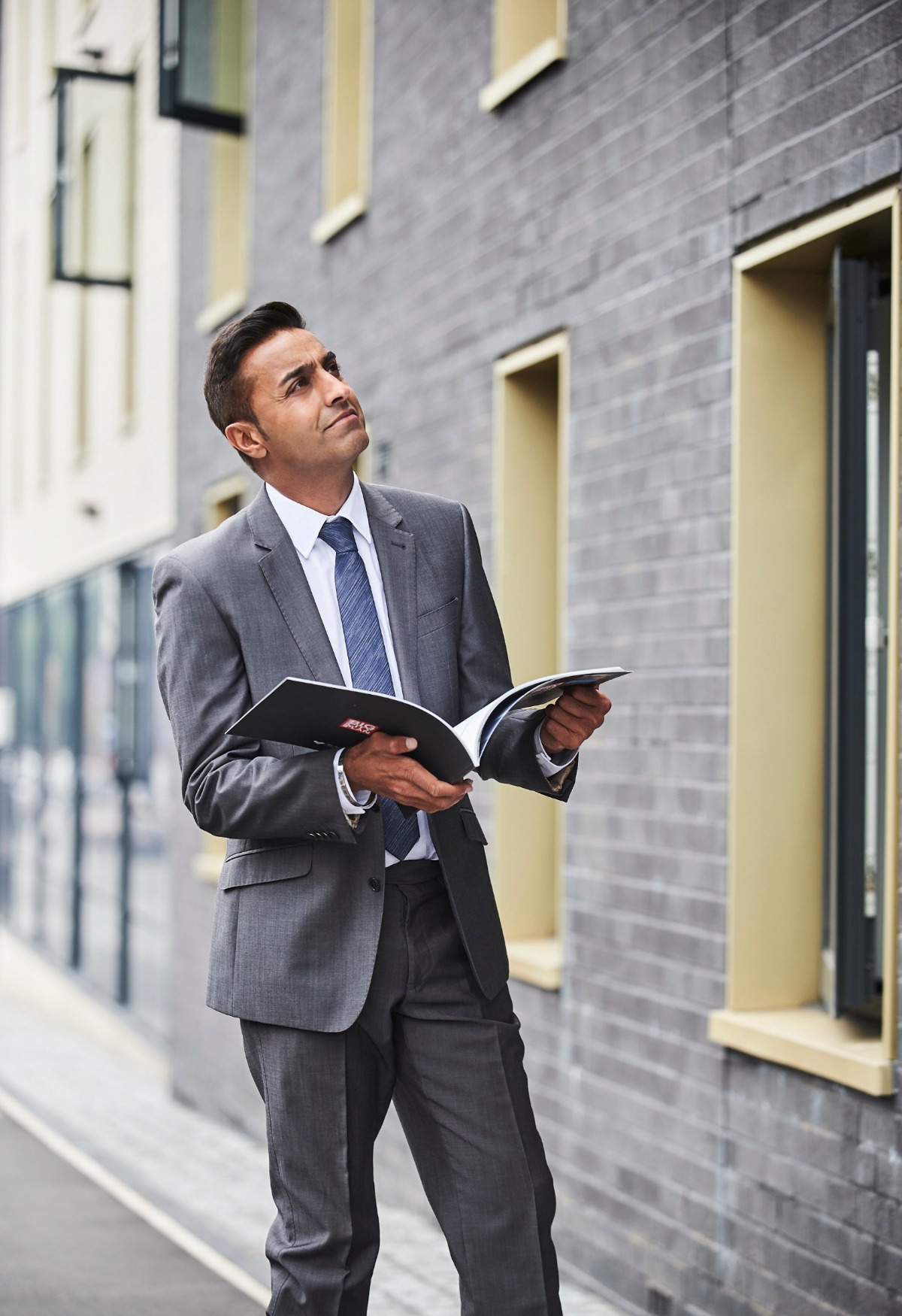 A professional man in a grey suit holding a brochure stands outdoors near a modern brick building.