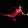 mohan-randhawa-14 A male football player in a bright red Liverpool kit performs an athletic leap against black background.