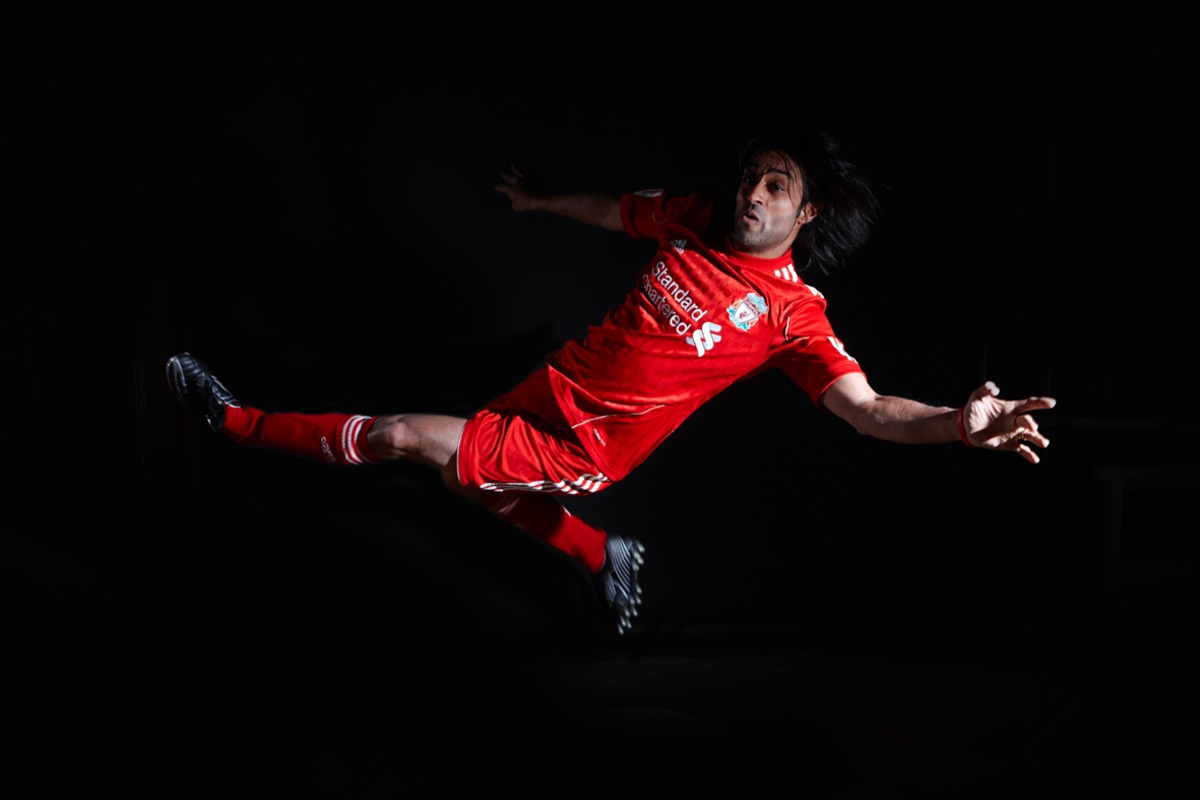 A male football player in a bright red Liverpool kit performs an athletic leap against black background.
