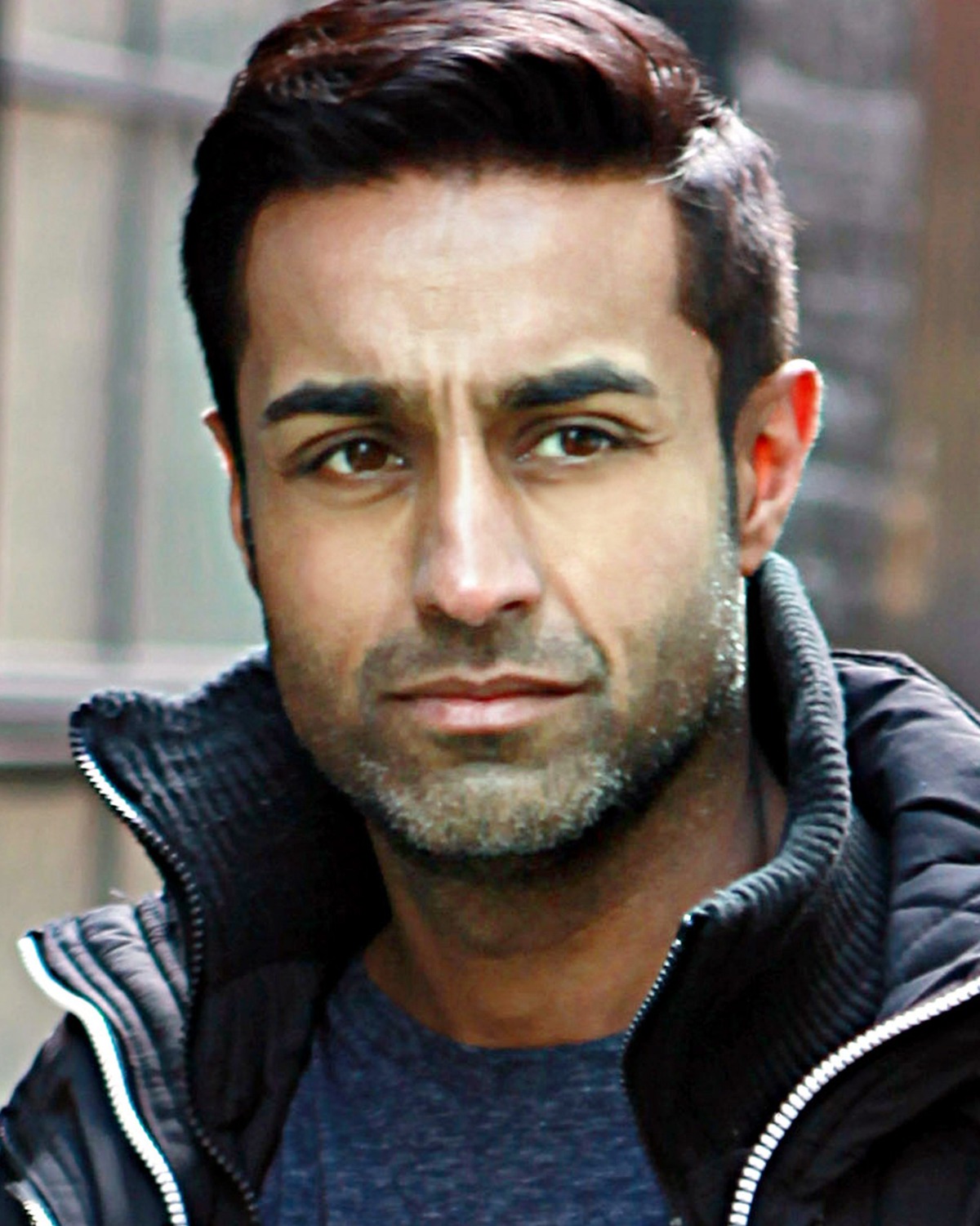 A focused man with dark hair and stubble wears a dark jacket in this crisp headshot portrait.