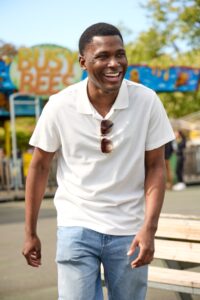 A smiling man wearing a white polo shirt walks outside at a bright, sunny local amusement park.