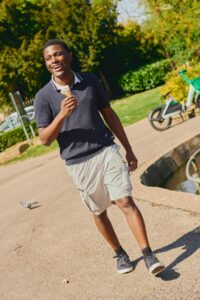 A smiling man enjoys an ice cream while walking through a bright and sunny public park.