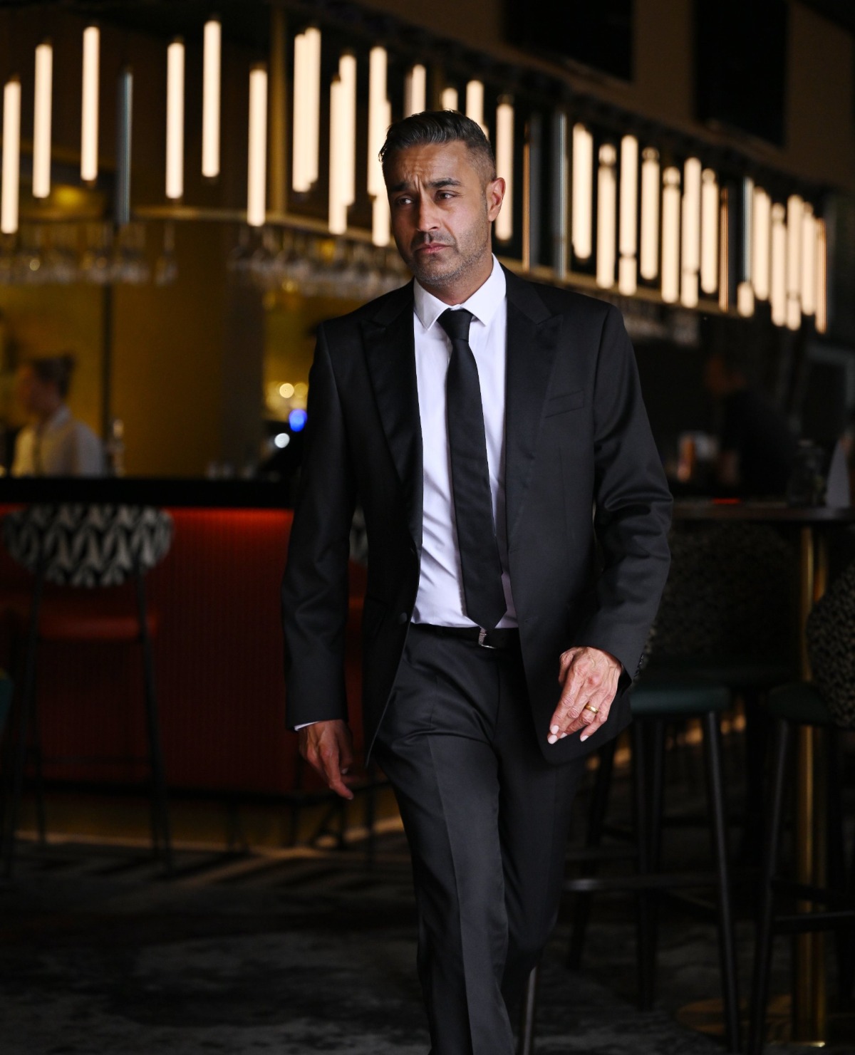 A man in a sharp black suit walking through a contemporary bar setting with warm interior lighting.
