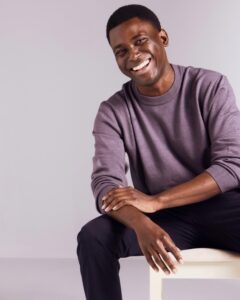 A smiling Black male model wears a casual purple sweater against a clean, neutral studio background.