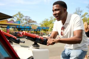 A man wearing a white polo shirt laughs joyfully while playing a game at an outdoor fairground.