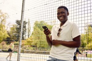 A smiling man using his mobile phone while standing by a tennis court on a sunny day.