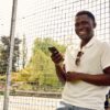 deondre-cumberbatch-10 A smiling man using his mobile phone while standing by a tennis court on a sunny day.